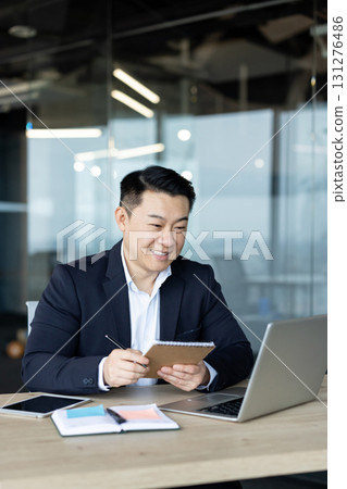Vertical photo of an Asian young man working in an office at a laptop, smiling at the screen and taking notes in a notebook. 131276486