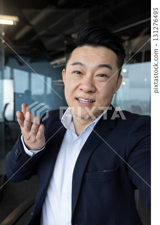 Vertical close-up photo of a young Asian man in a suit standing in an office and talking to a camera he is holding. Vertical close-up photo of a young Asian man in a suit standing in an office and talking to a camera he is holding. 131276495