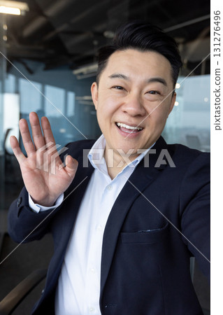 Vertical close-up photo of a young Asian businessman in the office, smiling and waving at the camera he is holding in his hand. Vertical close-up photo of a young Asian businessman in the office, smiling and waving at the camera he is holding in his hand. 131276496
