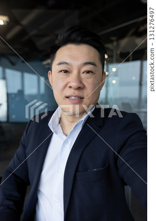 Vertical close-up photo of a young Asian man in a suit standing in an office and talking to a camera, he is holding in his hand. 131276497