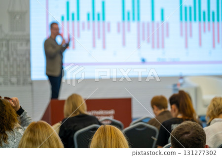 Business presenter on stage with large digital chart showing market trends during conference Business presenter on stage with large digital chart showing market trends during conference 131277302
