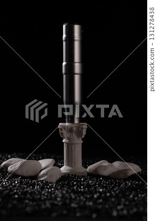 tattoo machine stands on a small white column with decorative details surrounded by smooth stones in a dark setting. This scene captures the artistry and tools of tattooing. tattoo machine stands on a small white column with decorative details surrounded by smooth stones in a dark setting. This scene captures the artistry and tools of tattooing. 131278438