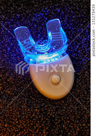 new teeth whitener device rests on textured black surface with bright blue LED light illuminating its transparent mouthpiece designed for home teeth whitening. 131278439