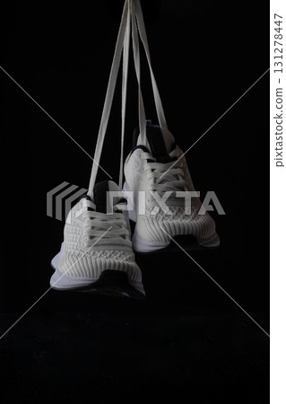White sneakers are suspended by their laces on a black backdrop highlighting their contemporary design and versatility for everyday wear. Perfect for casual outings or sporty looks. 131278447