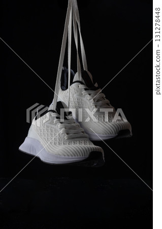 White sneakers hang from their laces against a solid black backdrop highlighting their stylish design and contemporary appeal. The focus is on the shoes and their details. 131278448