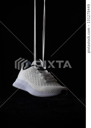 White sneakers dangle freely with shoelaces untied creating a striking contrast against a solid black background. The minimalist style highlights the clean lines and textures. White sneakers dangle freely with shoelaces untied creating a striking contrast against a solid black background. The minimalist style highlights the clean lines and textures. 131278449