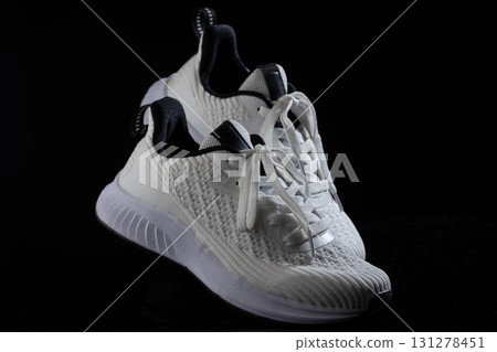 pair of white sneakers featuring a textured upper and black accents rests on a dark surface. The laces are untied adding a casual look. The sneakers highlight contemporary athletic footwear design. 131278451