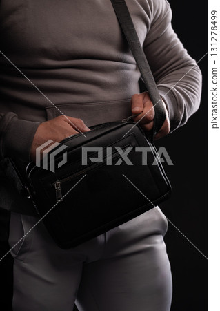 A man wearing a gray hoodie holds a black bag close to his body. The setting is dark emphasizing the man's stylish attire and the bag he carries. A man wearing a gray hoodie holds a black bag close to his body. The setting is dark emphasizing the man's stylish attire and the bag he carries. 131278499