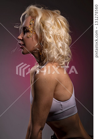 Blonde girl poses with gold glitter on her face showcasing her creative look against a vibrant background. This playful scene captures the joy of self expression and artistry. 131278560