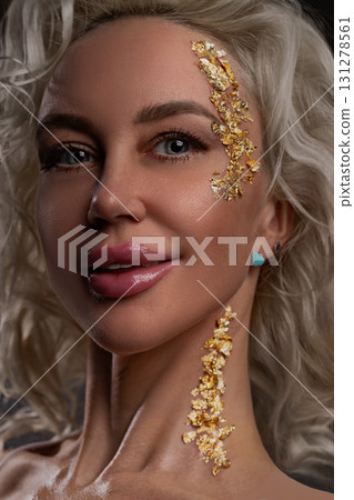 blonde girl is smiling and posing in a studio. Gold glitter decorates her face adding sparkle to her look. Her hair frames face beautifully complementing her confident expression. 131278561