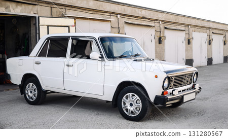 white classic car stands against the backdrop of a row of concrete garages with closed doors 131280567