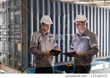 Two male workers in uniform and safety helmets checking documents and using a tablet in front of a shipping container, representing freight inspection, logistics coordination, and export operations. 131282475