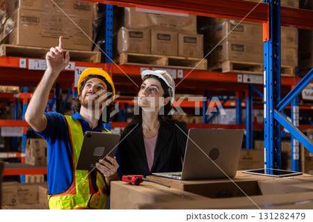 Two warehouse workers in safety helmets and vests using digital devices to inspect inventory and manage logistics inside a large storage facility with stacked cardboard boxes. 131282479