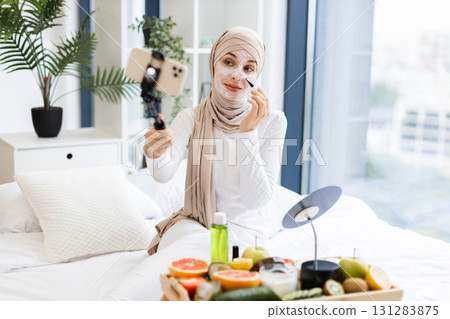 Young Muslim woman wearing headscarf applies face mask with brush while recording vlog showcasing skincare routine in bright and cozy room with natural light. Scene emphasizes self-care. 131283875