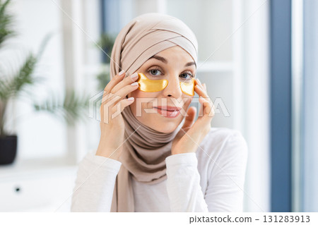 Young Muslim woman in beige hijab applying gold hydrogel eye patches for skincare in modern indoor environment. Promoting self-care and healthy lifestyle concept, model demonstrates beauty. Young Muslim woman in beige hijab applying gold hydrogel eye patches for skincare in modern indoor environment. Promoting self-care and healthy lifestyle concept, model demonstrates beauty. 131283913