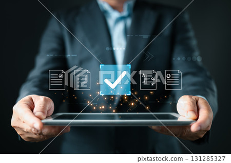 A business professional holds a tablet displaying floating document icons, a large checkmark symbolizing digital approve processes, focusing on business quality assurance and data management. A business professional holds a tablet displaying floating document icons, a large checkmark symbolizing digital approve processes, focusing on business quality assurance and data management. 131285327