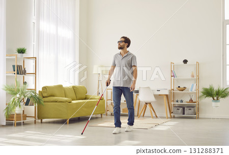 Bind young disabled man in casual clothes with cane walking in the living room at home. Bind young disabled man in casual clothes with cane walking in the living room at home. 131288371