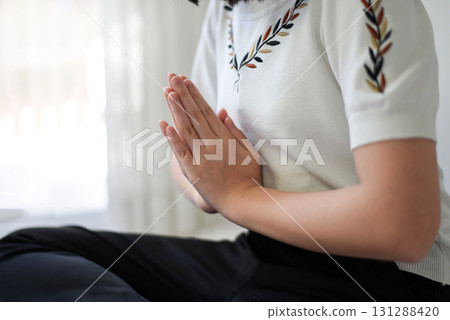 Close up image of woman's hands who is sitting cross-legged, hands folded, on the floor of the room at home To meditate to make the mind calm and relaxed. The room is bright with natural light. 131288420