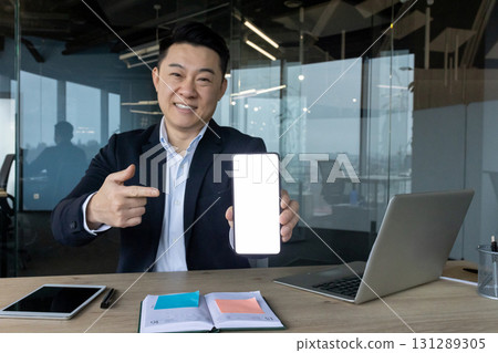 Portrait of a young smiling Asian man in a suit sitting in an office at a desk and pointing with his finger at the white background ,of the phone screen he is holding in his hands. 131289305