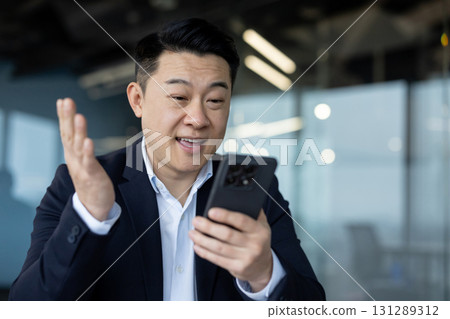 Close-up photo of a young Asian man in a suit sitting at a desk and talking on a video call via phone. 131289312
