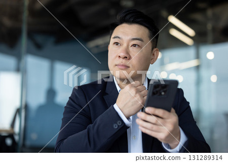 Close-up photo of a young Asian man in a suit sitting at his workplace with a phone in his hand, and looking out the window seriously. 131289314