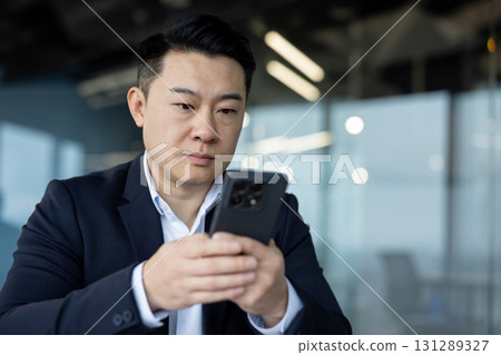 Close-up photo of a young focused and serious Asian businessman sitting in the office and using his mobile phone. Close-up photo of a young focused and serious Asian businessman sitting in the office and using his mobile phone. 131289327