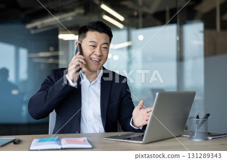 A young Asian man in a suit sits in the office in front of a laptop at a table and talks on a mobile phone, with a smile. A young Asian man in a suit sits in the office in front of a laptop at a table and talks on a mobile phone, with a smile. 131289343