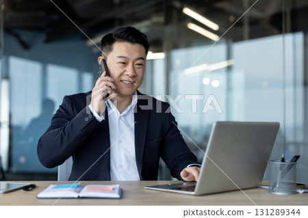 Smiling and successful young Asian man sitting in office, working on laptop and talking on mobile phone. Smiling and successful young Asian man sitting in office, working on laptop and talking on mobile phone. 131289344