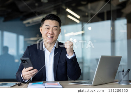 Portrait of a smiling young successful businessman sitting at a desk in the office, holding a mobile phone and showing a victory gesture with his hand. 131289360