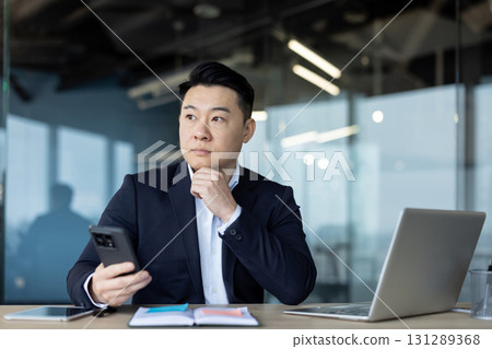 Serious young Asian male businessman sitting in the office at his desk, holding a phone in his hand and looking thoughtfully out the window. Serious young Asian male businessman sitting in the office at his desk, holding a phone in his hand and looking thoughtfully out the window. 131289368