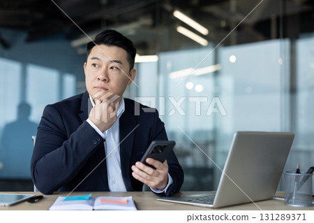 A serious young Asian male businessman in a business suit is sitting in the office at the desk, holding the phone in his hand and looking thoughtfully to the side. 131289371
