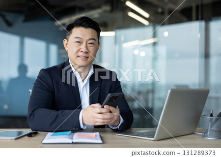 Portrait of a young smiling Asian businessman sitting at his desk in the office, holding a mobile phone and looking at the camera. Portrait of a young smiling Asian businessman sitting at his desk in the office, holding a mobile phone and looking at the camera. 131289373
