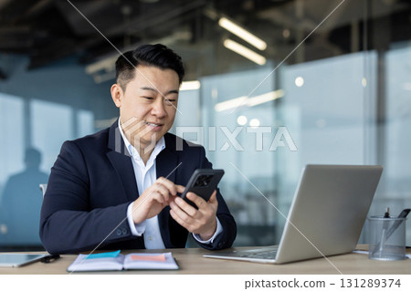 Smiling young Asian businessman sitting in office at desk with laptop and documents and using mobile phone. 131289374
