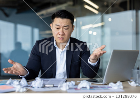 Upset young asian man in suit sitting in office at workplace and looking disappointed looking at crumpled paper on table, spreading hands. Upset young asian man in suit sitting in office at workplace and looking disappointed looking at crumpled paper on table, spreading hands. 131289376