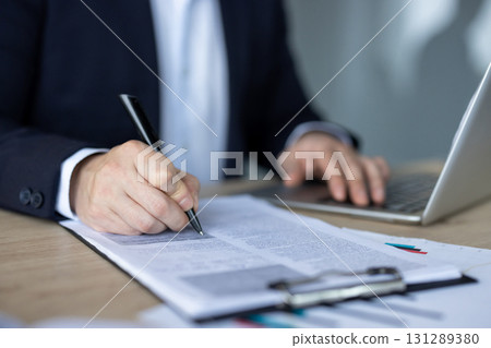Close-up photo of the hands, body parts of a young man in a suit who is writing documents with a pen at a table in the office, working with papers and a laptop. 131289380