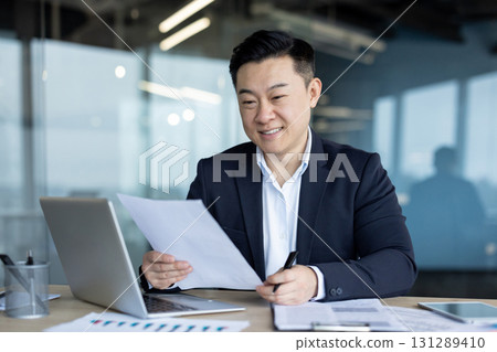 Smiling young Asian man in a suit working in the office at a desk with a laptop and reading documents and an agreement he is holding in his hands. 131289410
