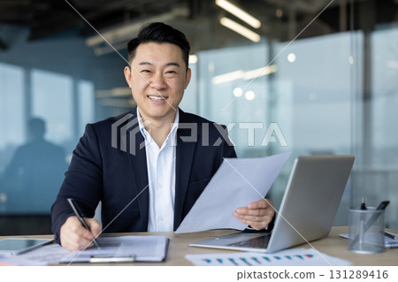 Portrait of a successful young Asian businessman working in the office with a laptop and documents, smiling and looking at the camera. 131289416