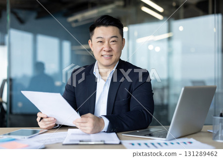 Portrait of a young successful and smiling Asian man in a suit sitting at a desk in the office, holding documents in his hands and looking at the camera. 131289462