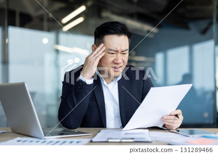 Upset young Asian man sitting at a desk in the office and holding his head while reading documents. Upset young Asian man sitting at a desk in the office and holding his head while reading documents. 131289466