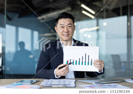 Portrait of a young smiling Asian man sitting in the office at a desk and showing documents with company graphs and revenues to the camera. Portrait of a young smiling Asian man sitting in the office at a desk and showing documents with company graphs and revenues to the camera. 131290189