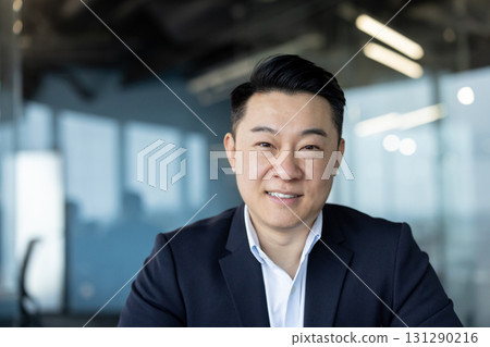 Close-up portrait of a young Asian businessman in a suit sitting at his desk in the office and smiling at the camera. Close-up portrait of a young Asian businessman in a suit sitting at his desk in the office and smiling at the camera. 131290216