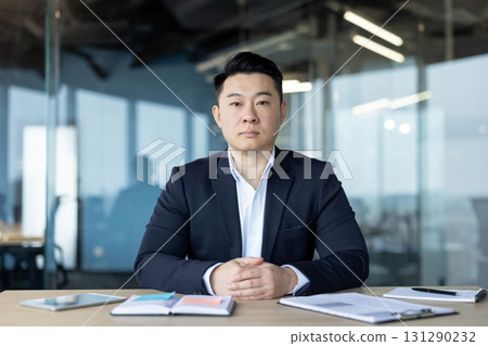 Portrait of a serious young Asian businessman man in a business suit sitting at a desk in the office and looking confidently at the camera. Portrait of a serious young Asian businessman man in a business suit sitting at a desk in the office and looking confidently at the camera. 131290232