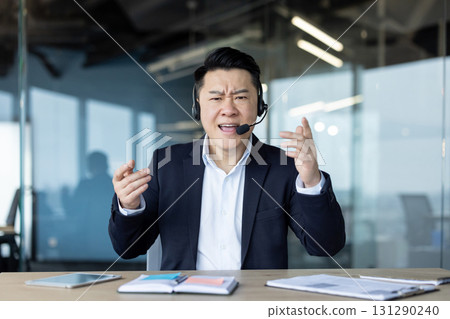 Portrait of a young Asian angry man in a suit and headset sitting at a desk and talking emotionally to the camera. 131290240