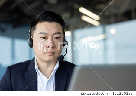 Close-up photo of a young serious Asian man in a suit sitting in an office at a workplace and working on a laptop, with concentration. Close-up photo of a young serious Asian man in a suit sitting in an office at a workplace and working on a laptop, with concentration. 131290252