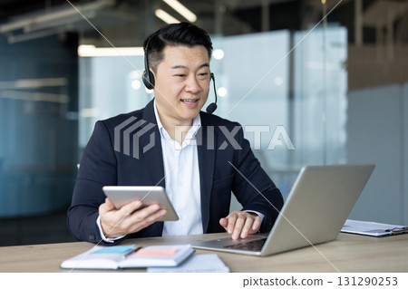A businessman wearing a headset engages in a video call while working on a laptop and tablet, smiling at the camera. A businessman wearing a headset engages in a video call while working on a laptop and tablet, smiling at the camera. 131290253