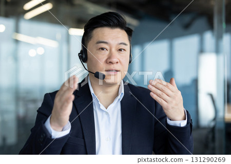 Close-up portrait of a young Asian man in a suit and headset talking to the camera and gesturing with his hands. Close-up portrait of a young Asian man in a suit and headset talking to the camera and gesturing with his hands. 131290269