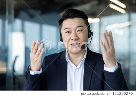 Close-up portrait of a young Asian man in a suit and jacket standing in the office, talking and gesturing with his hands to the camera. Close-up portrait of a young Asian man in a suit and jacket standing in the office, talking and gesturing with his hands to the camera. 131290270