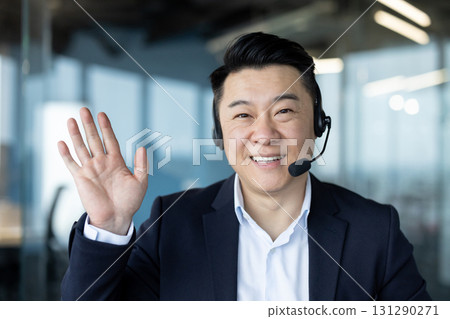 Close-up portrait of a smiling young Asian man in a suit and headset sitting in an office facing the camera, greeting and waving. Close-up portrait of a smiling young Asian man in a suit and headset sitting in an office facing the camera, greeting and waving. 131290271