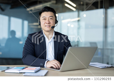 Portrait of a smiling young Asian man in a suit and headset working in an office with a laptop and looking at the camera. Portrait of a smiling young Asian man in a suit and headset working in an office with a laptop and looking at the camera. 131290293