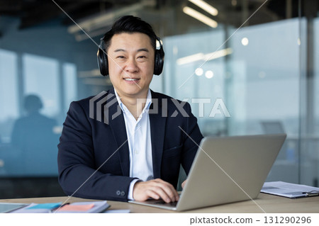 Portrait of a confident and confident Asian man in a suit and headphones sitting in the office at a laptop, and looking at the camera with a smile. Portrait of a confident and confident Asian man in a suit and headphones sitting in the office at a laptop, and looking at the camera with a smile. 131290296